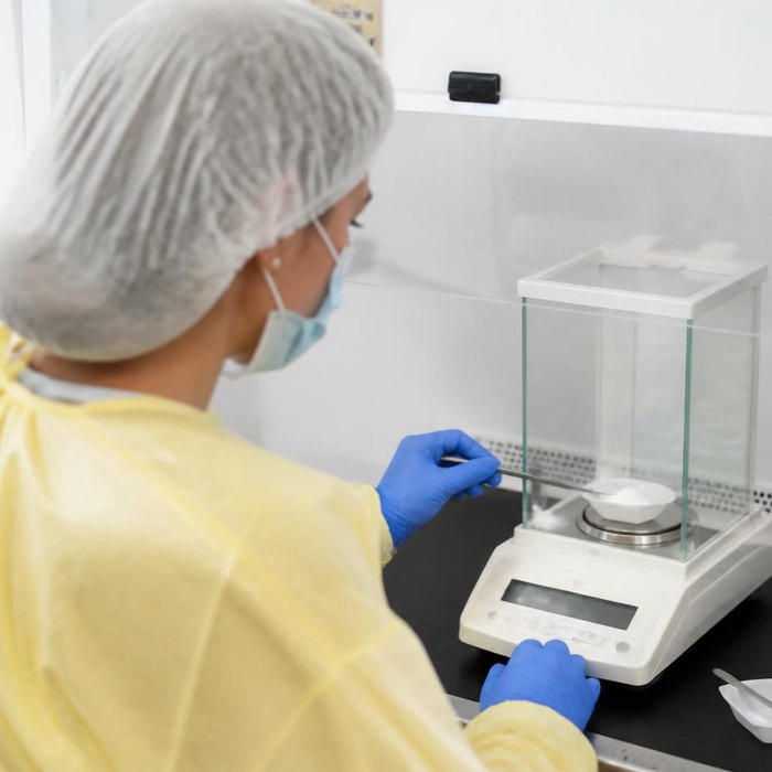 Pharmacist in a protective gown weighing powder on an analytical balance inside a containment hood (illustrative image)