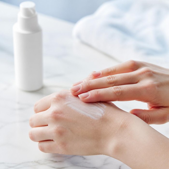Hands rubbing white cream into the back of one hand, with a plain pump bottle behind them (illustrative image)